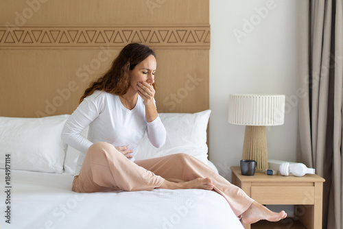 Adult woman sitting on bed covering mouth, feeling nauseous at home bedroom. Health discomfort concept, lifestyle realism, morning sickness, illness awareness, and personal wellbeing moment