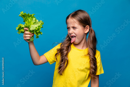 Photography Little cute kid girl in yellow t-shirt making grimaces, showing tongue and refusing to eat healthy fresh lettuce salad on blue background