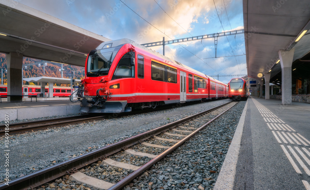 Fototapeta premium Red modern passenger train on mountain railway station in Swiss Alps in autumn. High-speed train, railway platform and cloudy sky. Bernina Express. Rail transport. Station in St. Moritz, Switzerland