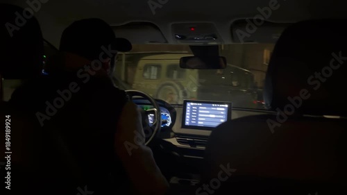 Rear view of a man driving a car at night in the city. Silhouette of a male driver holding the steering wheel with a glowing modern dashboard and infotainment screen. Urban night traffic concept.