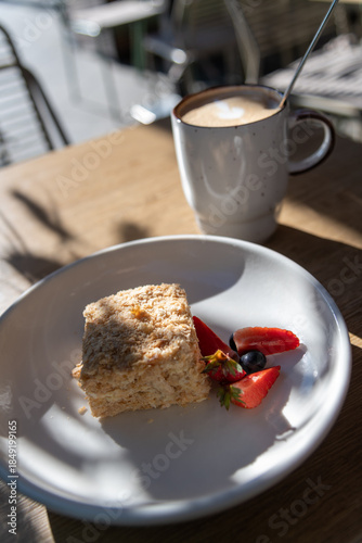 Cake sits on a white plate next to fresh strawberries and blueberries. A cup of coffee stands nearby. Sunlight shines through, creating shadows on the wooden table.