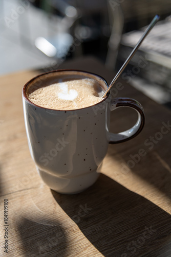 A ceramic cup filled with coffee sits on a wooden table. Sunlight casts a shadow as a spoon rests inside the mug. Surrounding elements are blurred in the background.