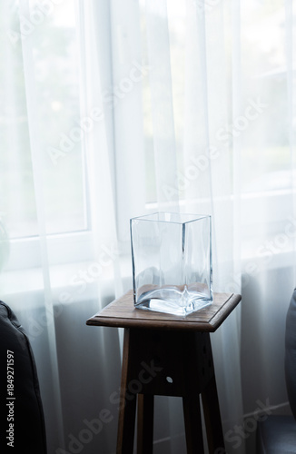 Empty glass vase on wooden stool near window in minimalist interior