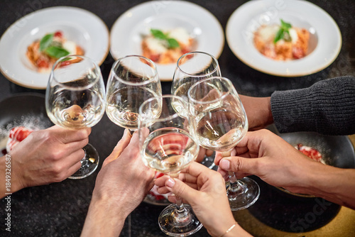 Diverse group of young hands clinking wine glasses during celebratory meal, visible plates of food in background, multiethnic gathering sharing toast