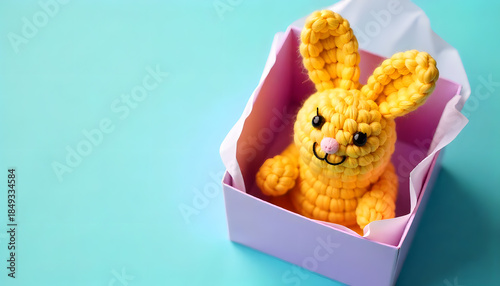Cute handmade crocheted bunny toy sitting inside a pastel paper box on a light blue background. Playful and minimal studio composition with soft colors and large copy space. easter gift concept, ad