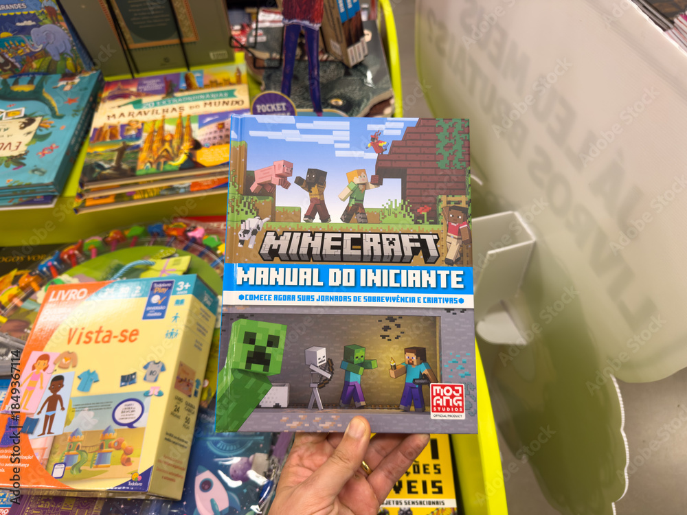 Fototapeta premium BELO HORIZONTE, MG, BRAZIL, DECEMBER 12, 2025 A person's hand holds a vibrant Minecraft Beginner's Manual book, showcasing popular gaming culture in a bright bookstore aisle.
