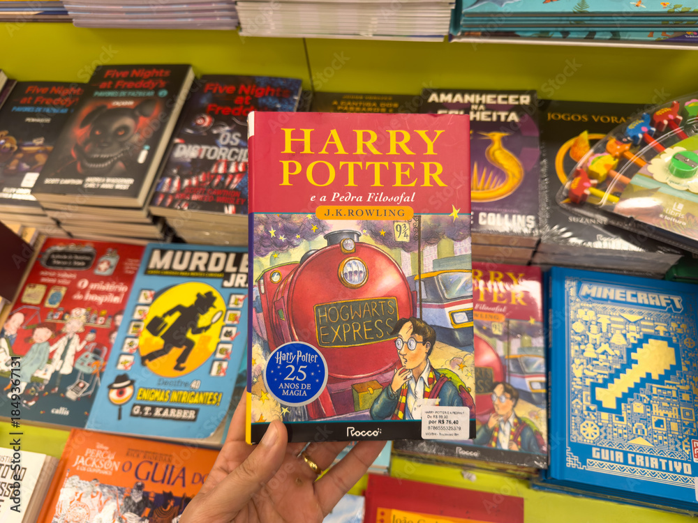 Naklejka premium BELO HORIZONTE, MG, BRAZIL, DECEMBER 12, 2025 Hand holding a special edition of Harry Potter and the Philosopher's Stone in a bustling bookstore, celebrating 25 years of magic.