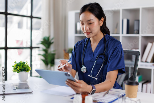 Quadro su tela An attractive and professional Asian female doctor with blue coat or Asian female doctor working on her medical cases at her desk in the office at a hospital