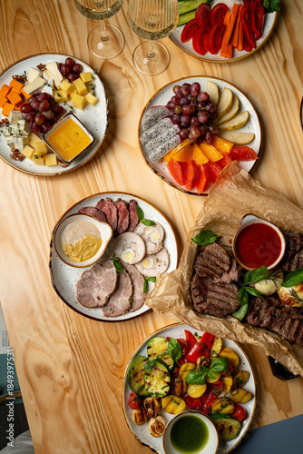 At a wooden table, a colorful spread of gourmet cheeses, fresh fruits, roasted meats, and assorted veggies awaits, perfect for sharing and enjoying together