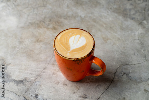 A rich latte presented in a bright orange mug showcases delicate heart-shaped art on its frothy surface. The warm drink sits gently on a cool gray table, inviting enjoyment