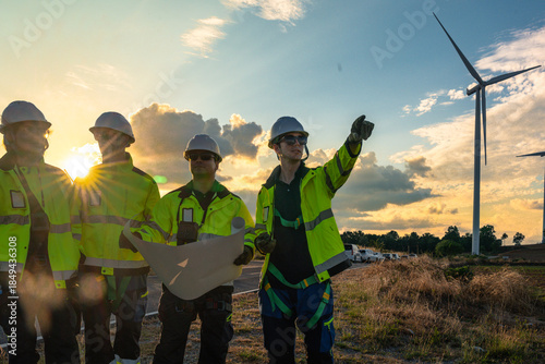Fotografie Team of four engineers in white hard hats and high-visibility gear pointing at wind turbines during sunset