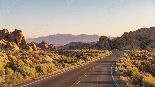 A scenic asphalt road winds through an arid desert landscape during golden hour. Distant mountains are visible under a clear sky. The scene is bathed