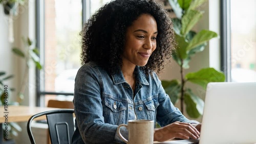 A happy Black woman with curly hair smiles while typing on a laptop at a cafe. She wears a denim jacket. A mug and plant are nearby