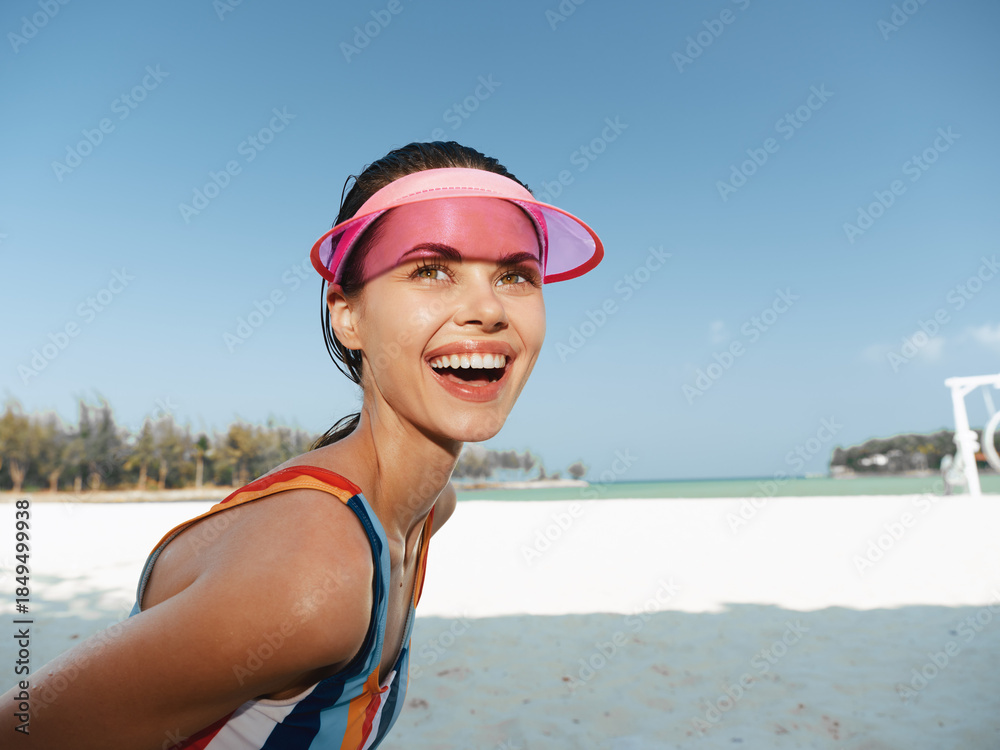 Φωτογραφία Joyful woman on a sunny beach, wearing a colorful swimsuit and pink visor, smiles toward the camera as white sand and turquoise water frame her energetic pose