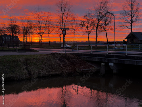 A sunset in a little village in the Netherlands with a channel in the foreground with reflection of the sky