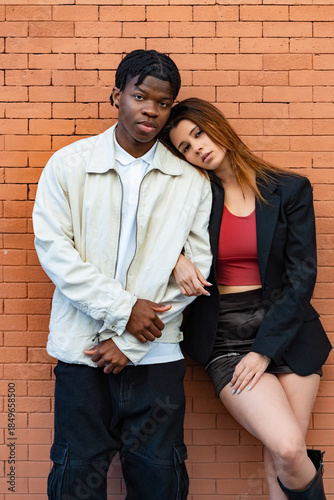 Fashionable young multi ethnic couple posing together leaning against a brick wall, embracing and looking serious