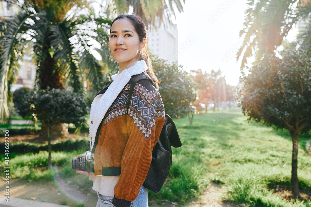 Obraz premium Woman with camera and backpack in a park, wearing a jacket and scarf, enjoying outdoor sunlight and nature during daytime, ready for street photography and exploration