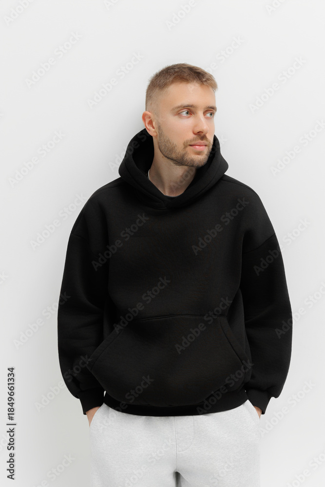 Obraz premium Man stands with hands in pockets wearing a black hoodie in a simple setting with a plain background