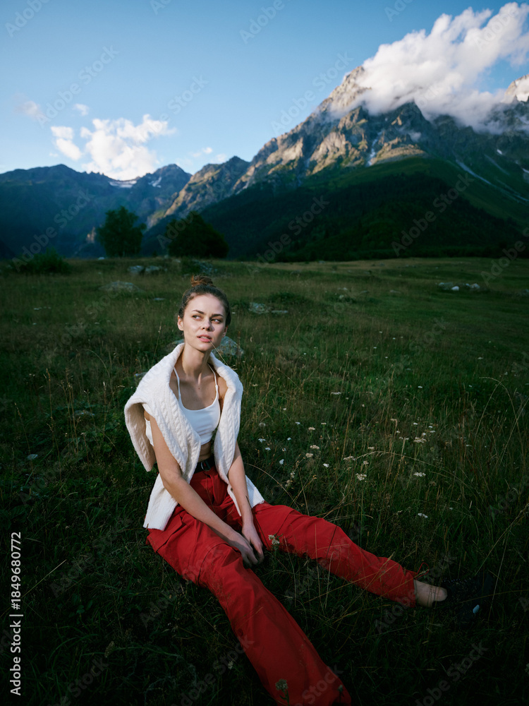 Fototapeta premium Woman sitting in a meadow with red pants and white top, rugged mountains in the background, cloudy sky, outdoor fashion mood, serene landscape and nature inspired scene