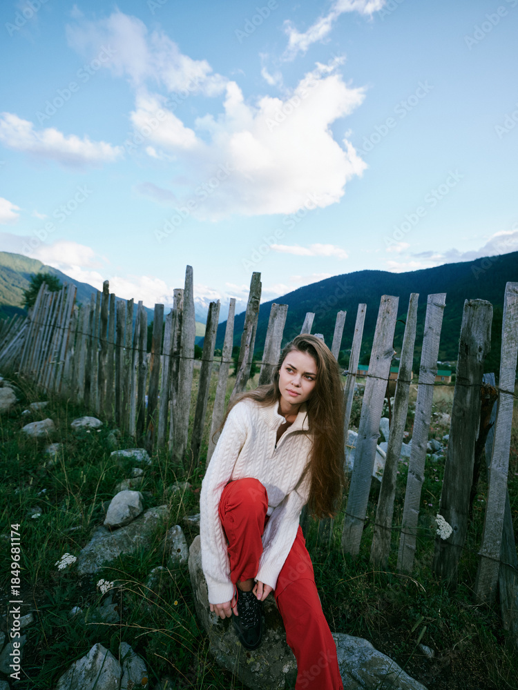 Obraz premium Woman in cozy sweater and red pants sits by a rustic fence in a green hillside landscape, capturing a quiet countryside mood for lifestyle, travel and outdoor photography