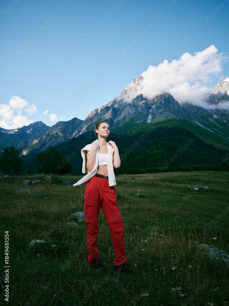 Fototapeta premium Woman in red pants stands in a wide meadow with towering mountains, bright sky, and a light jacket, conveying bold adventure and freedom in nature. Her confident pose suggests exploration, fitness,