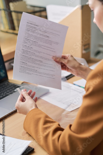 Caucasian young adult woman reviewing printed documents at desk, holding paper in one hand and sheet protector in other, working near open laptop in modern office environment, focusing on paperwork