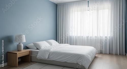 A modern bedroom with a large bed, white bedding, and a wooden nightstand. Soft blue walls and sheer curtains allow natural light to enter the room.
