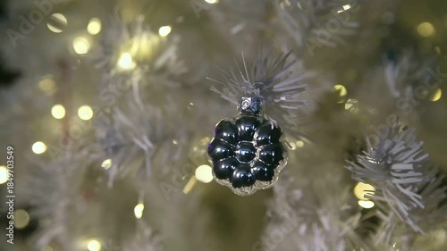 A close-up of a black ornament hanging on a white Christmas tree. The tree is adorned with warm white lights, creating a festive atmosphere.