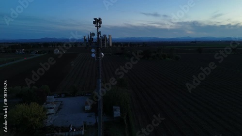 Aerial footage of a tall telecommunication tower equipped with antennas and satellite dishes, transmitting signals against a clear dark blue sky at dusk in a rural landscape