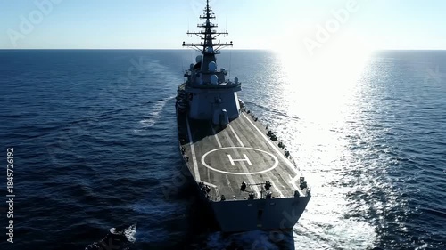 Aerial view of a military warship sailing through the ocean at sunrise with a helipad on deck