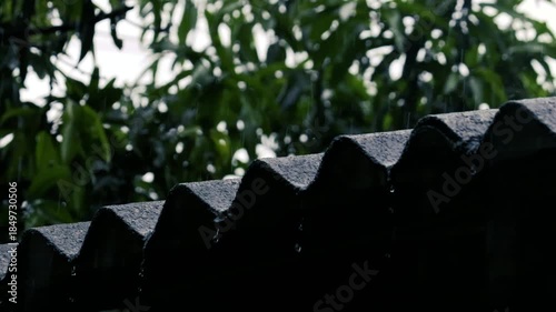 heavy rain pouring on dark corrugated roof silhouette with blurred green trees in background
