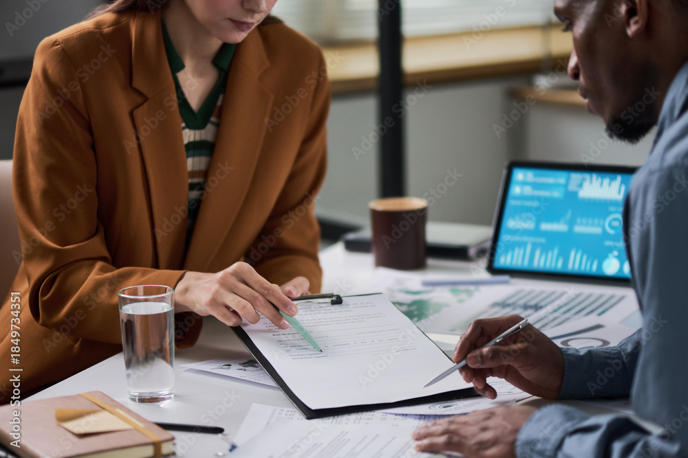 Obraz premium Caucasian young adult woman and Black young adult man reviewing financial documents together at desk, both focusing on paperwork with digital tablet displaying charts in background