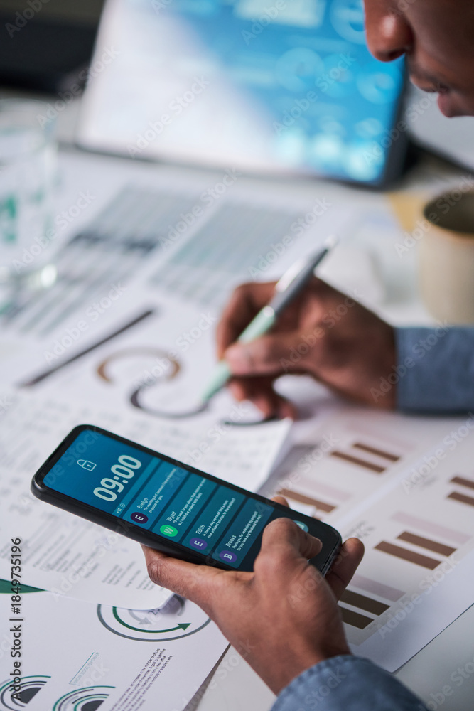 Fototapeta premium Young adult Black man holding smartphone with notifications visible while analyzing printed charts and graphs at desk, multitasking with pen in hand and digital device in workspace