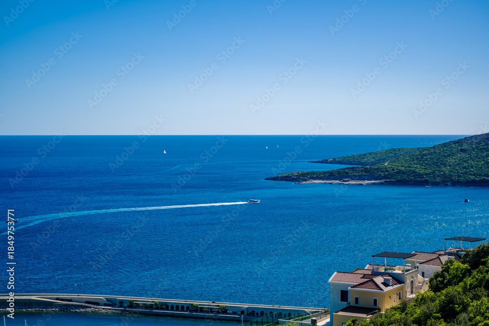 Obraz premium A scenic view of the Adriatic Sea in Montenegro. Clear blue waters stretch to the horizon, with a green hillside and coastal houses visible.