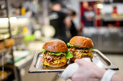 Two gourmet burgers on a metal serving tray in a commercial kitchen setting. Hand holding tray with motion blur, focus on food