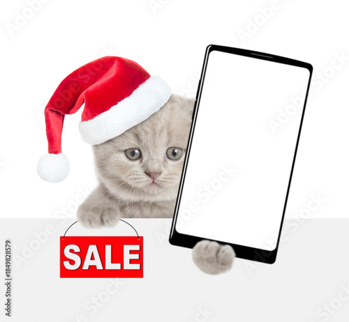 Cute kitten wearing red christmas hat holds smartphone with white blank screen in it paw looks above blank white banner. and showing signboard with labeled 