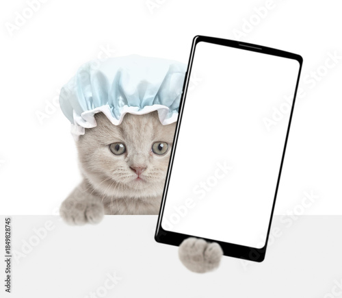 Funny tiny kitten wearing shower cap holds big smartphone with white blank screen in it paw above blank white banner. isolated on white background