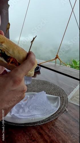 Handheld motion eating a sub sandwich on a wooden balcony with the view of foggy forest in the distance, candid authentic moment 
