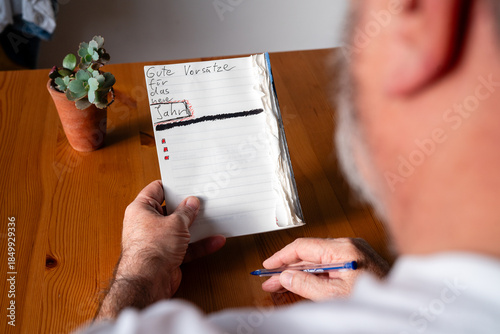 Ein älterer Mann sitzt an einem Holztisch und hält ein offenes Notizbuch mit der Überschrift 