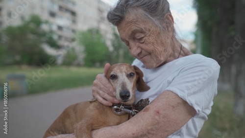 Senior 90-year-old woman with gray hair and deep wrinkles sits outdoors In assisted living facility on bench with small dachshund dog. Old female hugs and cuddles pet in the park on a bench.