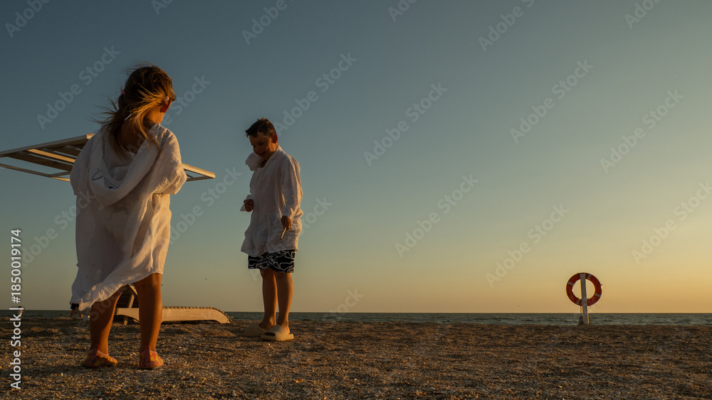 Fototapeta premium Two children, a boy and a girl, standing on a beach at sunset, wearing white cover-ups after swimming, with a tranquil sea and clear sky providing a relaxed atmosphere during their vacation.