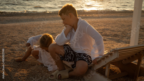 Two young children, a boy and a girl, relax on a peaceful beach, contemplating the warm golden sunset light reflecting on the sea, experiencing a quiet summer evening moment.