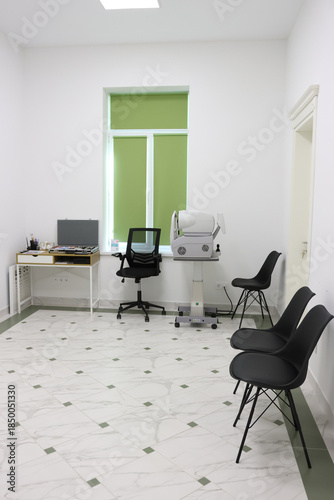 Interior of an ophthalmologist's office with a slit lamp biomicroscope, a table with a laptop and two chairs.