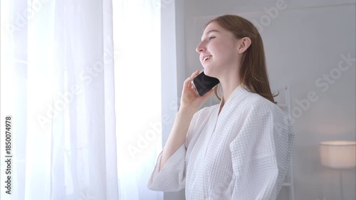 A young woman and a new morning with use cell phone to talk in the morning sunlight by the window of the house before taking a shower and going to work