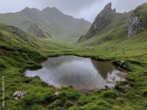 Misty mountain scene featuring a tranquil pond, green grass, and rocky peaks under overcast skies