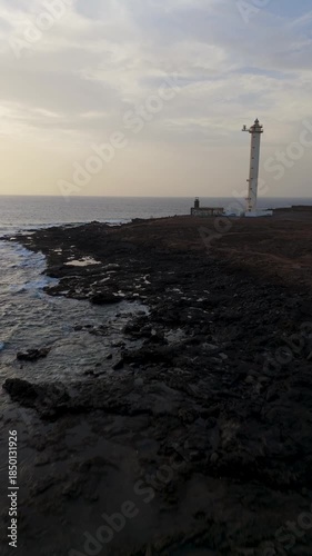 Vertical Drone Flight Past Faro de Pechiguera Lighthouse at Golden Hour, featuring the Rugged Volcanic Coastline of Lanzarote, Canary Islands.