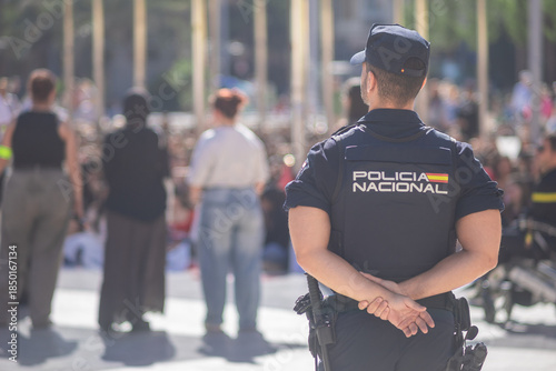 A back view of a Spanish National Police officer standing guard at a sunlit public event in Valencia. The blurred crowd and urban background emphasize a focus on public safety and security.