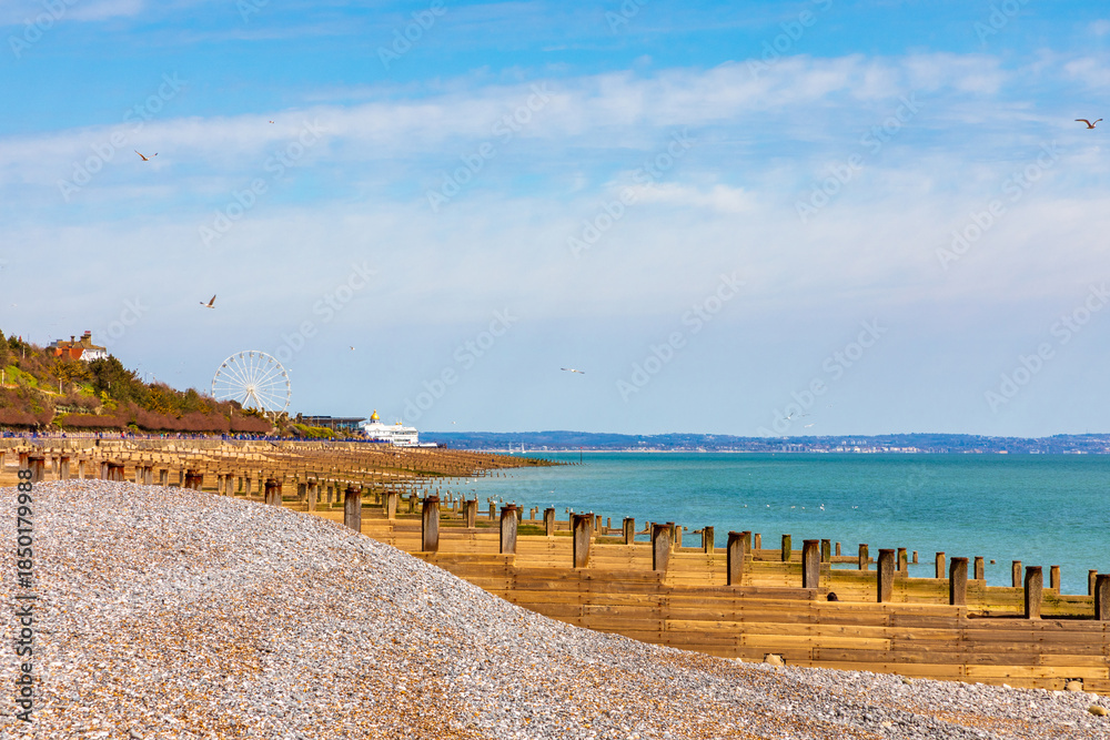 Fototapeta premium Eastbourn, Coastal beach scene with pebble shore, wooden groynes, and ferris wheel in the distance