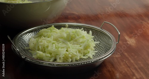 View back on the peel the zucchini. 4K close-up: A chef transfers sliced zucchini, but first squeezes the juice out of the zucchini and places it in a metal bowl on a wooden plate set on a rustic