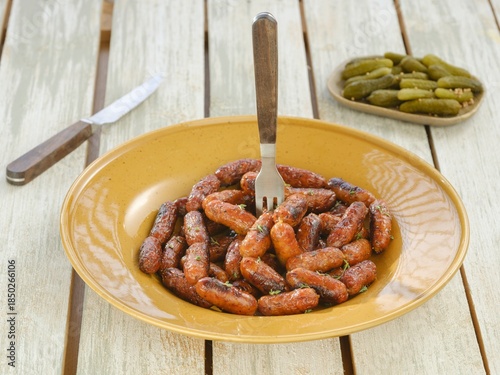 Fotografía A tantalizing array of small grilled sausages sits on a yellow plate, accompanied by a dish of petite pickles